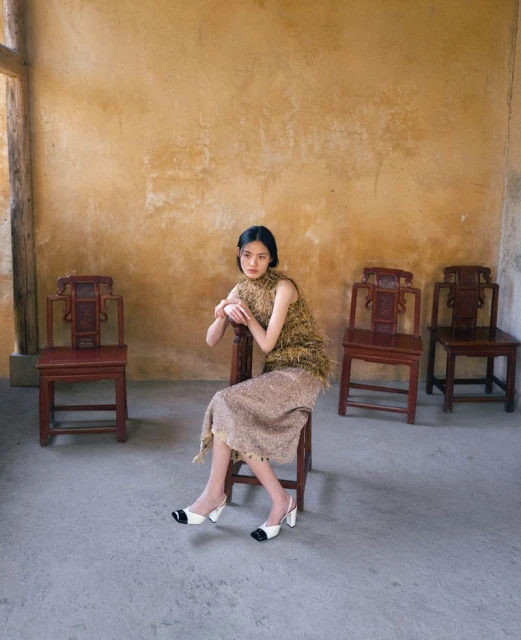 A woman sits on a wooden chair in a room with tan walls, wearing a sleeveless, shaggy dress and white-and-black heels, shot in soft, diffused light.