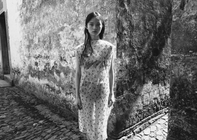 Monochrome portrait of a young Asian woman in floral dress standing in an alleyway with textured walls and cobblestone ground.