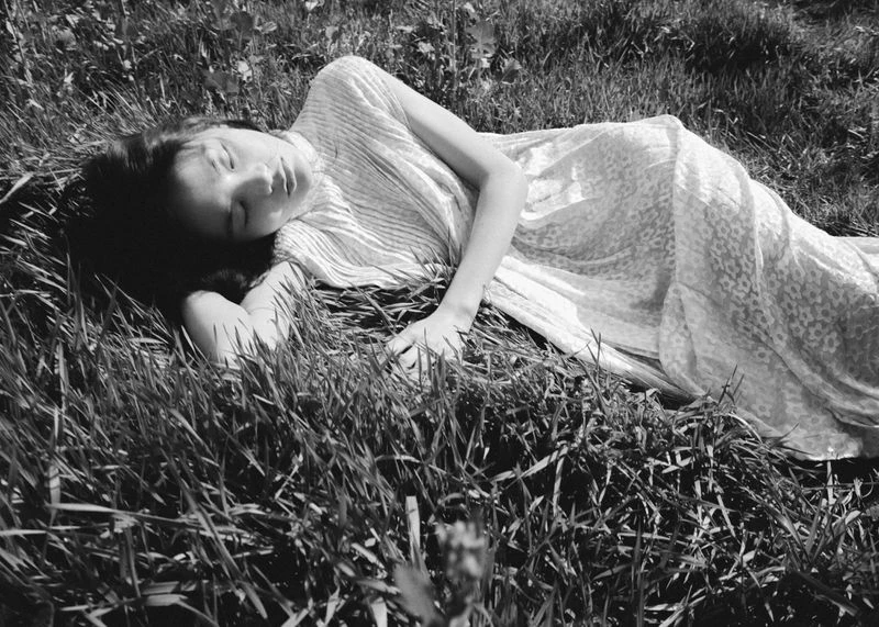 An adult female is lying on her back in a field of tall green grass, wearing a patterned white dress, eyes closed, in a black-and-white photograph.