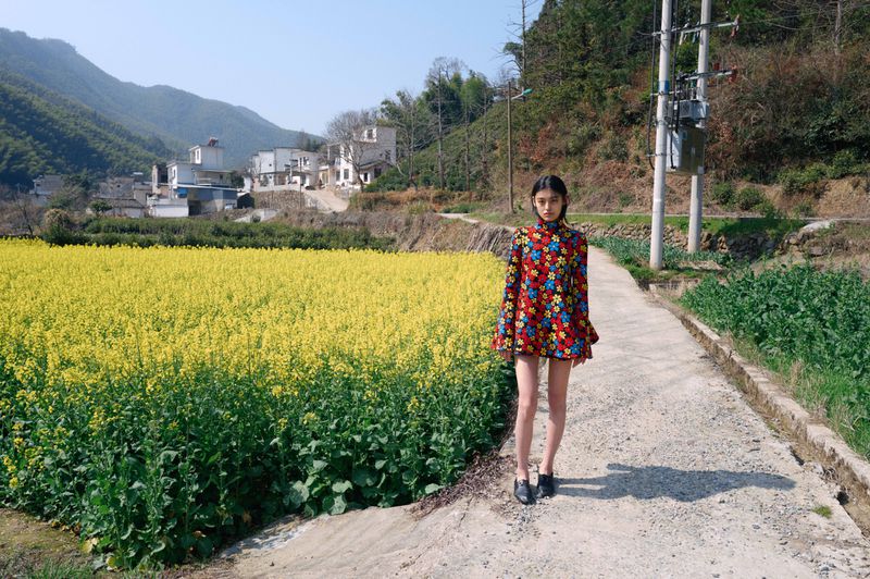 A woman stands on a rural road in a floral dress near a field of yellow flowers on a sunny day, with mountains and buildings in the background.