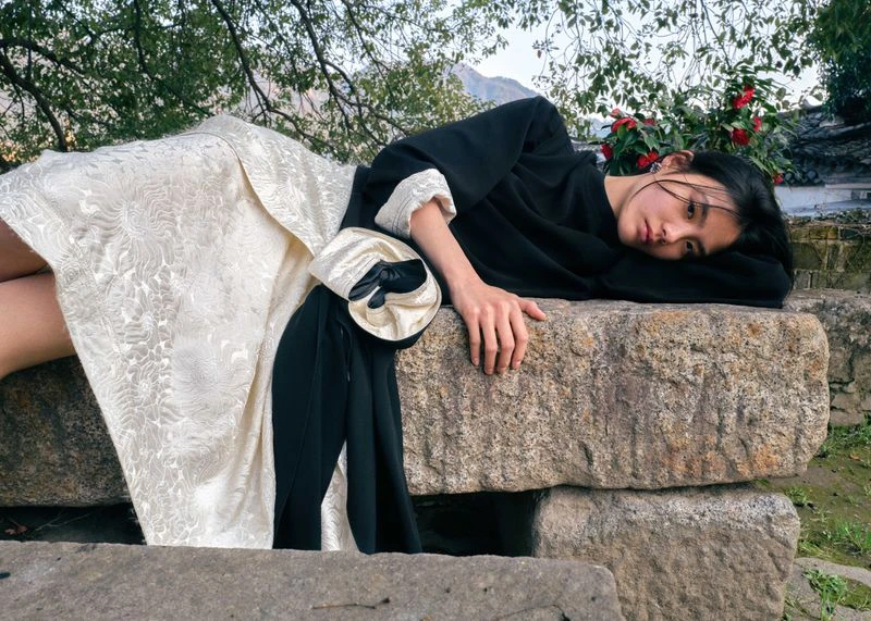 Asian woman in a black top and patterned skirt lying on stone blocks outdoors with a red-flowered bush, trees, and building in background.