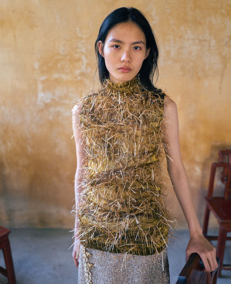 A woman with dark hair is posing in a sleeveless top made of tan straw-like material and a light-colored skirt, inside a room.