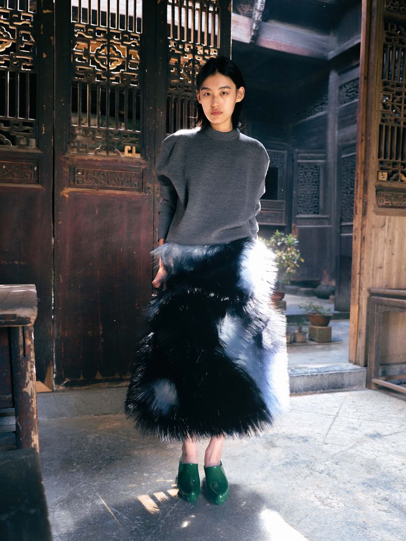 A woman wearing a gray sweater and a long black and white fur skirt stands in a traditional wooden building, looking at the camera.