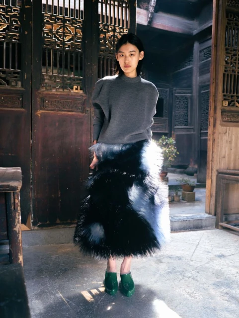 A woman wearing a gray sweater and a long black and white fur skirt stands in a traditional wooden building, looking at the camera.