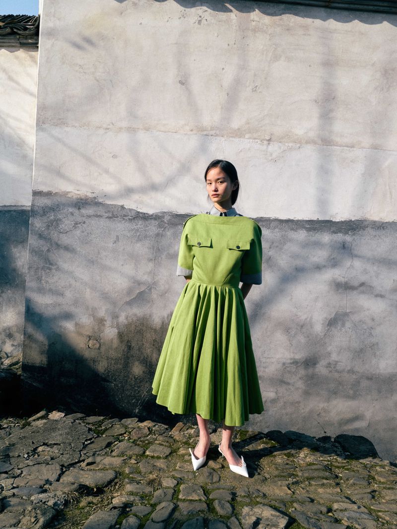 Model posing in a bright green short-sleeved pleated midi dress and white pointed heels on a cobblestone path.