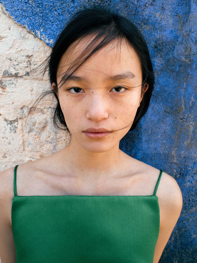 Close-up portrait of an Asian woman with dark hair and green dress against a textured blue and white painted background.