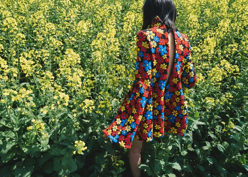 A woman stands in a field of yellow flowers wearing a black dress with a colorful floral pattern. The open back is visible.