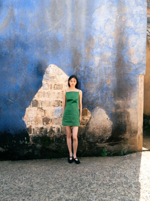 Young woman in a green dress stands against a textured blue wall, fashion shot, outdoor setting with natural light and concrete ground.