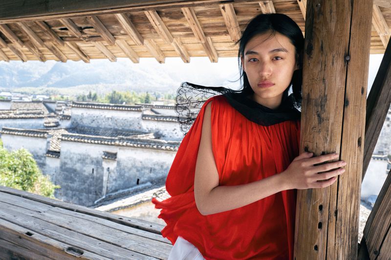 Asian woman in red top poses against wooden pillar with village view in background, showcasing fashion and architectural elements.