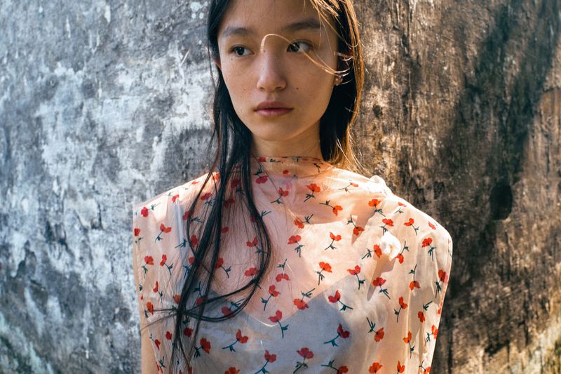 Portrait of a young woman with long dark hair wearing a floral-patterned top, posing against a textured wall in natural sunlight.