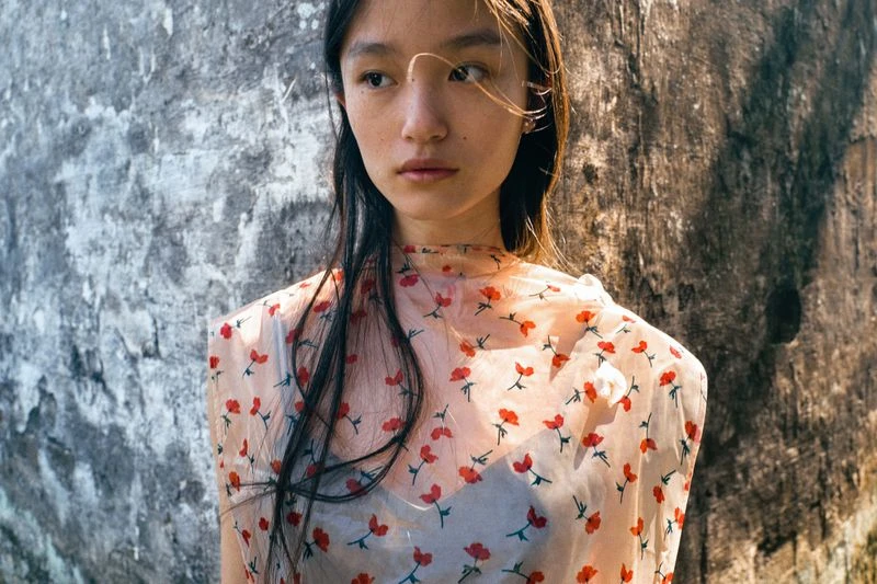 Portrait of a young woman with long dark hair wearing a floral-patterned top, posing against a textured wall in natural sunlight.