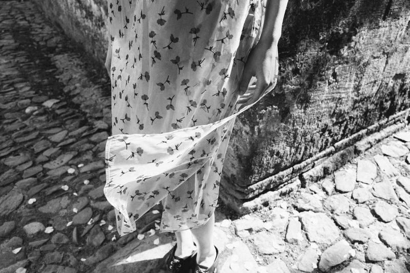 Woman in floral dress stands on cobblestone street next to a stone wall in a black and white photograph, capturing fashion and architecture.