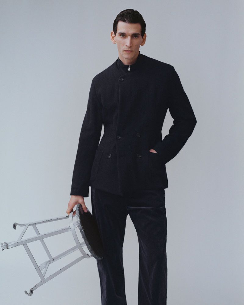 A stylish adult male poses in a photo studio wearing a modern black jacket and matching pants while holding a metal stool.