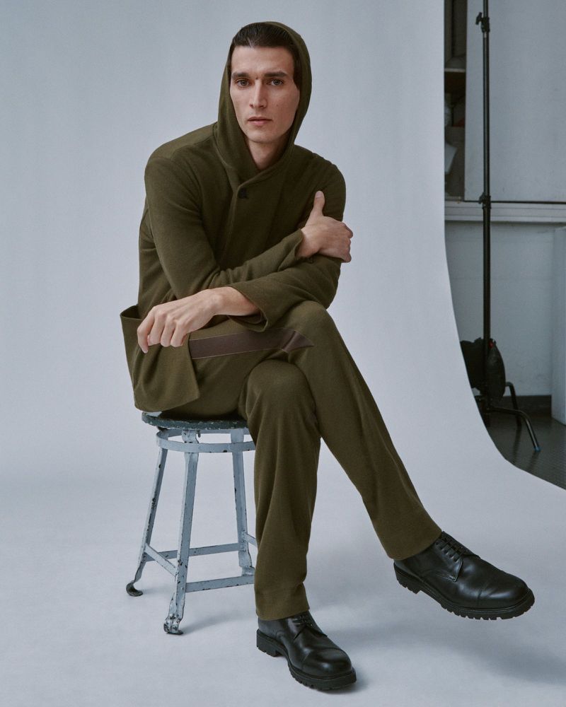 A man in an olive green hooded top and matching trousers sits on a metal stool against a white studio seamless backdrop.
