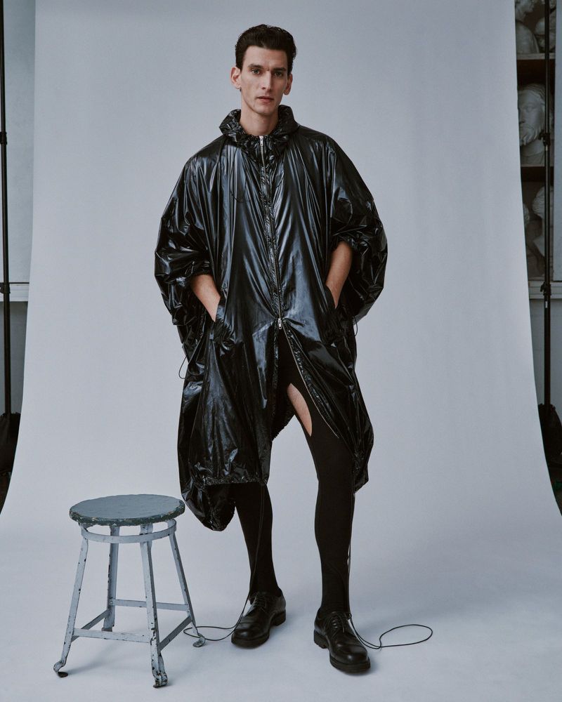 Male model posing in a glossy black hooded poncho with black tights and shoes in a neutral studio environment with a grey stool.