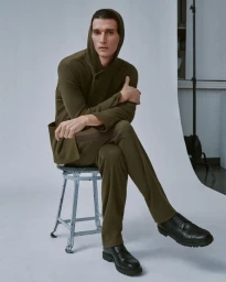 A man in an olive green hooded top and matching trousers sits on a metal stool against a white studio seamless backdrop.