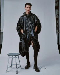 Thibaud Charon wearing a glossy black hooded poncho and tights next to a metal stool against a white seamless backdrop.