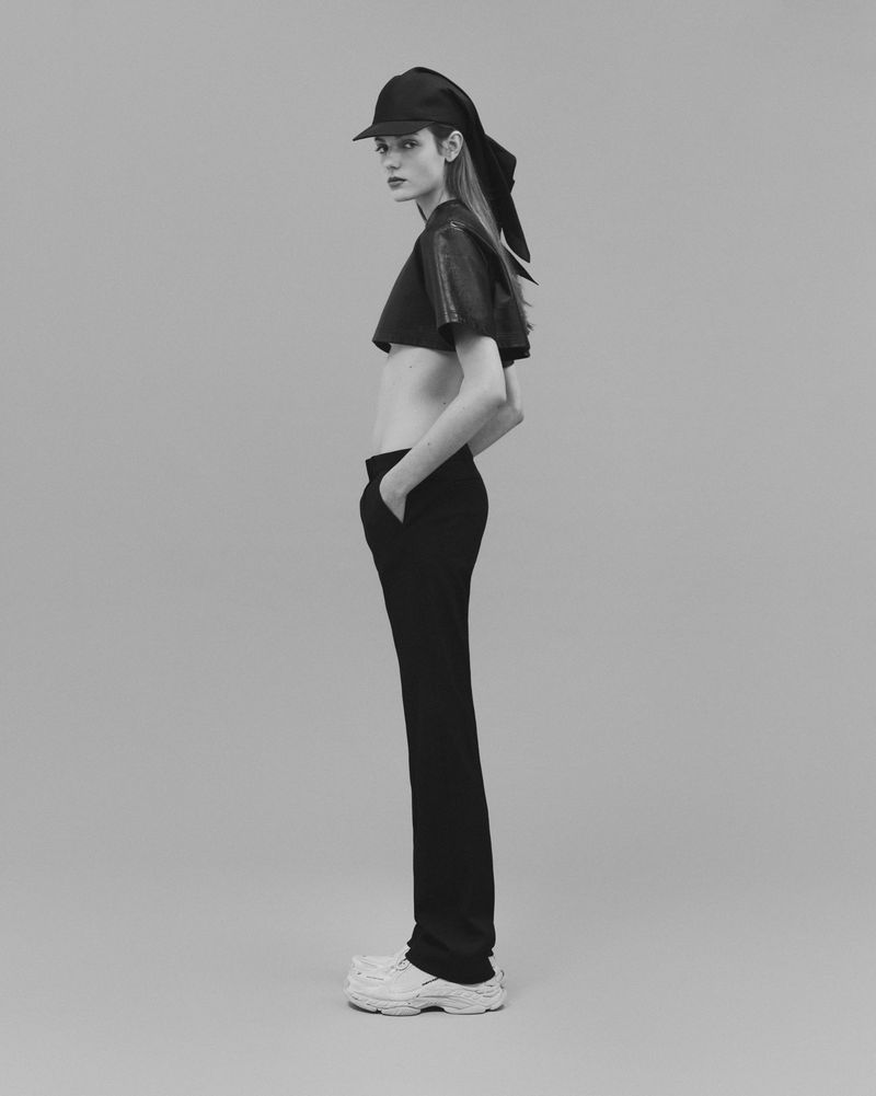 Monochrome studio shot of a young adult female fashion model in a cap, crop top, and trousers posing in profile.