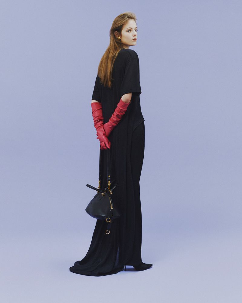 Fashion shot of an adult woman in a black dress and red gloves, standing in front of a lilac background, holding a black handbag.