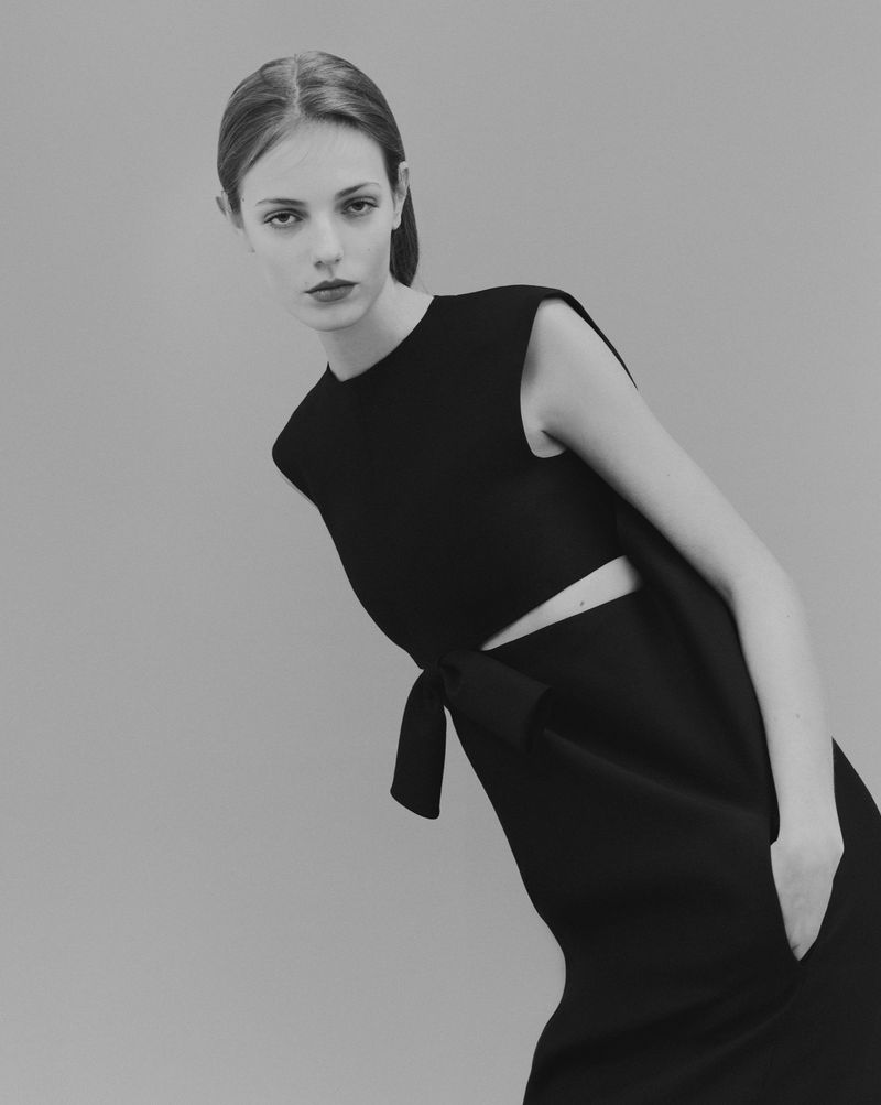 Monochrome fashion shot of a slender, fair-skinned adult female model in a stylish black dress with a tied waist, posing against a gray backdrop.