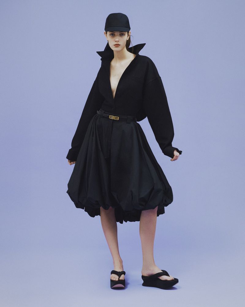 A woman in a black outfit with a plunging top, voluminous skirt, cap, and platform sandals, poses against a light purple background.