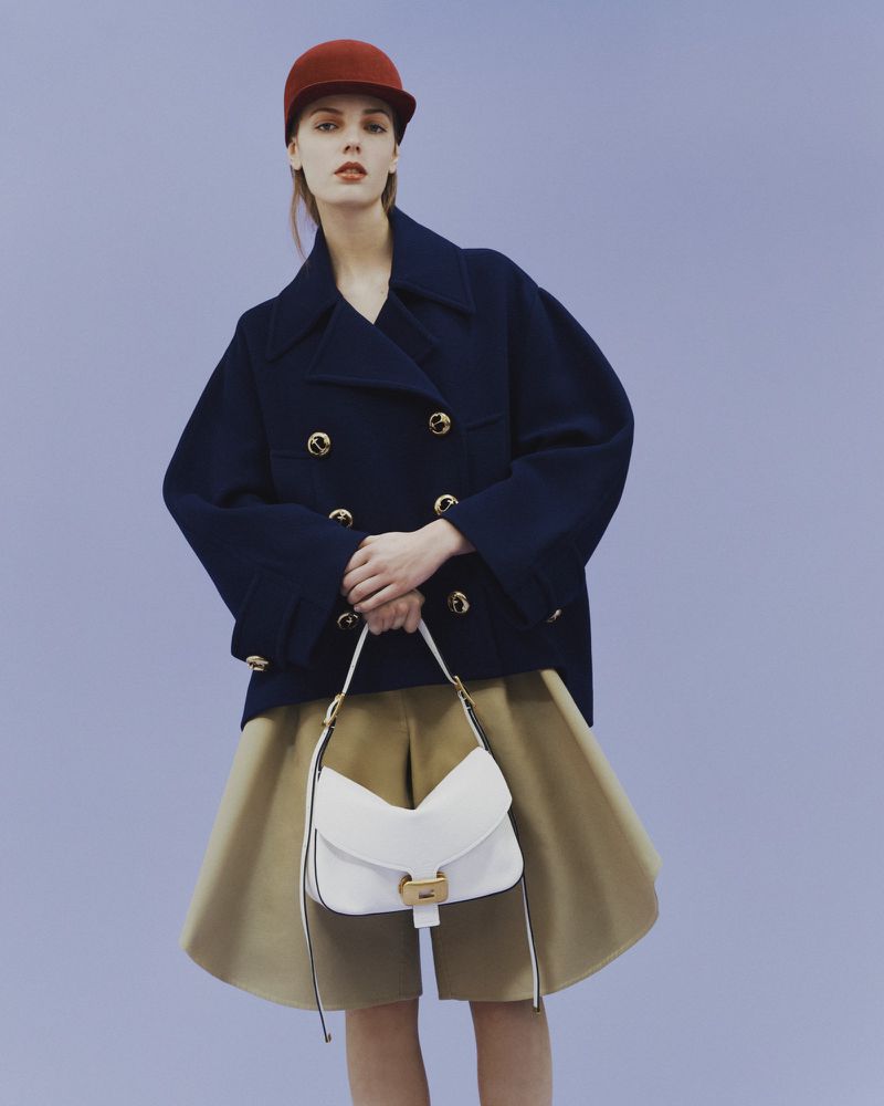 A young female model poses in a blue double-breasted jacket, tan shorts, and a rust-colored hat, holding a white handbag against a blue backdrop.