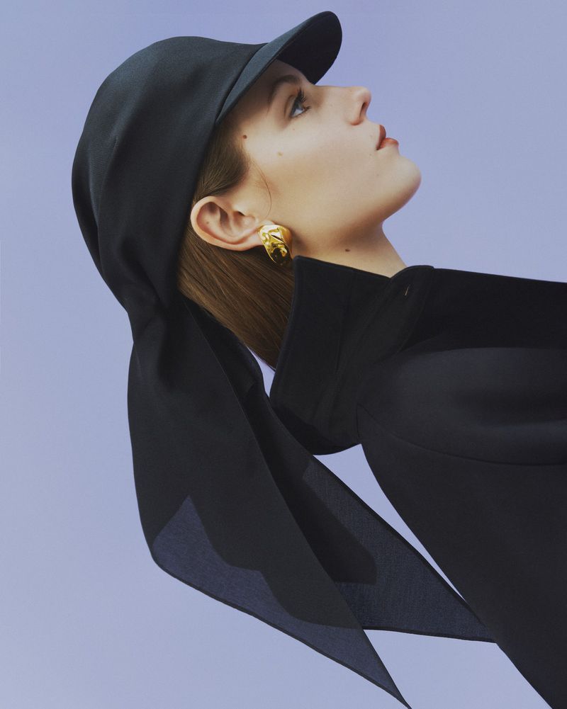 Woman in black cap and jacket poses against a light purple background, showcasing a fashion-forward look with focus on contemporary style.