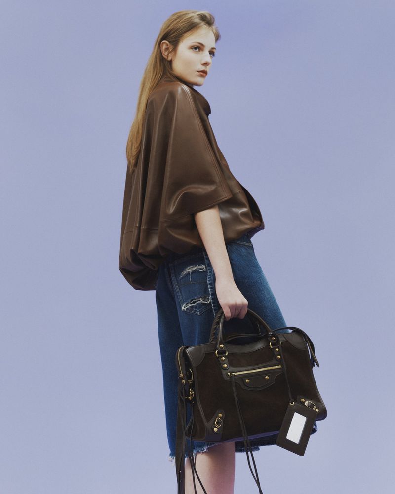 Model Matilde de Nard in a brown leather top and distressed denim holding a brown handbag against a lavender studio background.