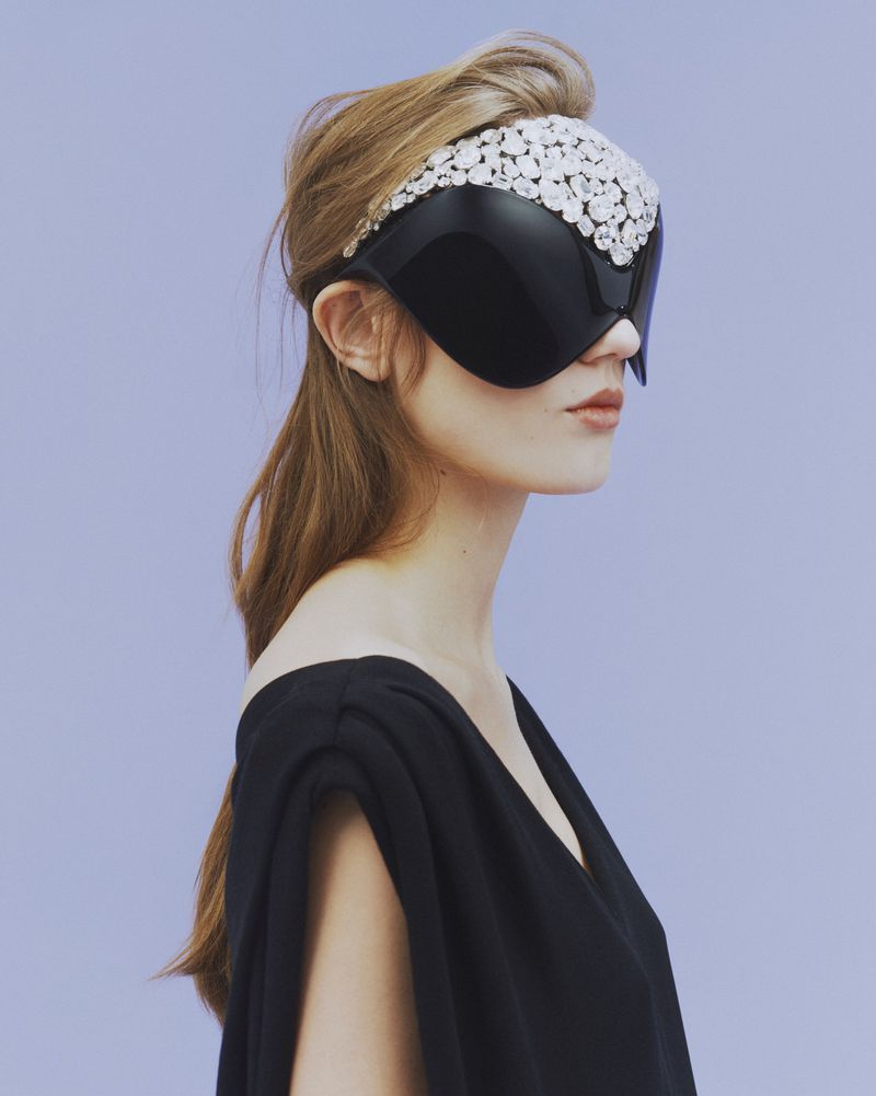 Profile of a woman wearing an avant-garde black visor with white beaded details against a solid lilac background in a studio setting.