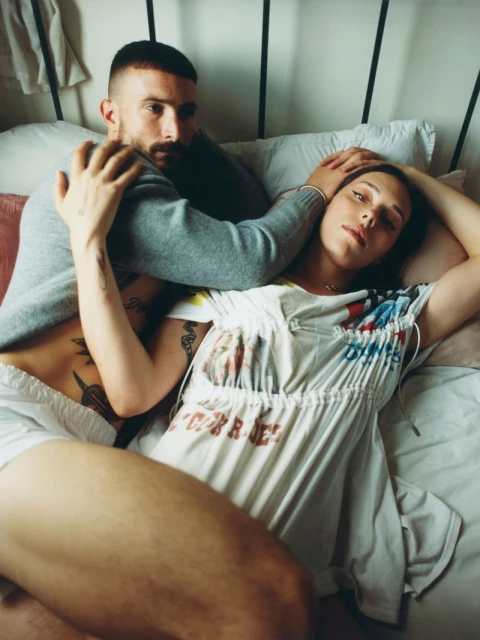 Intimate portrait of a couple in bed, the man with a beard and the woman in a printed t-shirt dress, soft lighting, indoor setting.
