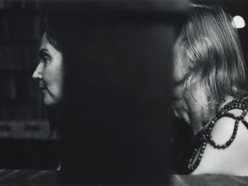 Monochrome portrait of two adult women, one in profile, showcasing beads and blurred features in a dark, indoor setting.