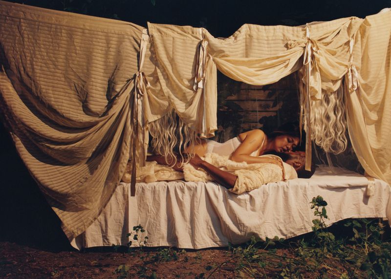 Two people, a man and a woman, are lying on a bed with a draped canopy outdoors, appearing relaxed in a dark, natural setting.