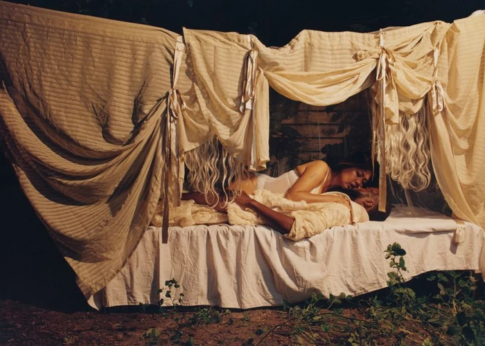 Two people lying together on an outdoor bed draped with hanging fabric sheets and curtains under dim lighting.