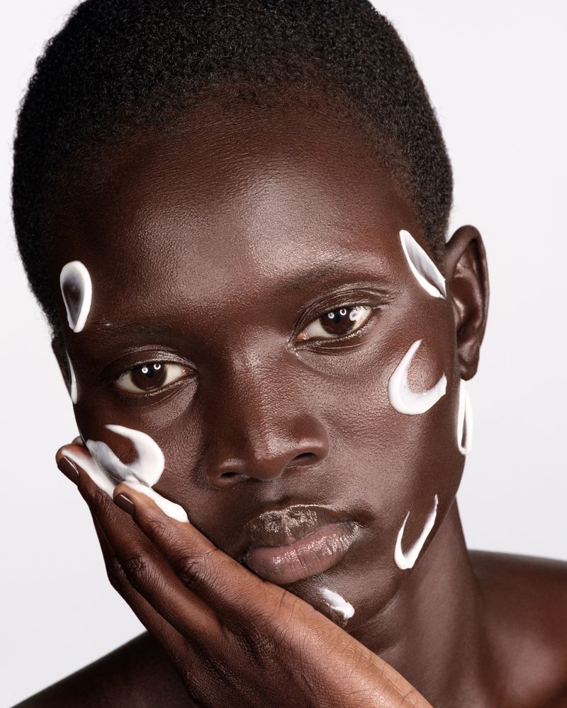 Close-up beauty portrait of a Black woman with short hair and white facial cream swatches against a white studio backdrop.