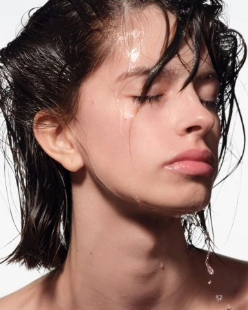 Close-up portrait of a young adult woman with wet dark hair and water droplets on her face against a clean white background.