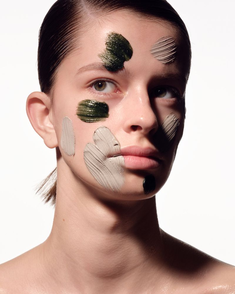 Close-up of a young woman's face with patches of different colored facial mask, set against a bright white background.