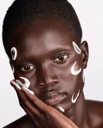 Close-up beauty portrait of a Black woman with short hair and white facial cream swatches against a white studio backdrop.
