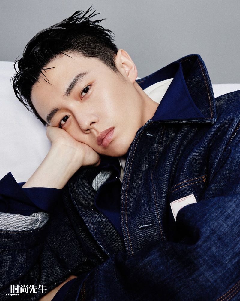 Portrait of a relaxed adult male with dark hair, wearing a denim jacket, resting his head on his hand in a casual indoor setting.