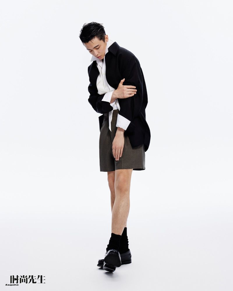 Fashionable Asian man in a black blazer and shorts stands in a studio with a slight downward gaze in a full-body shot.