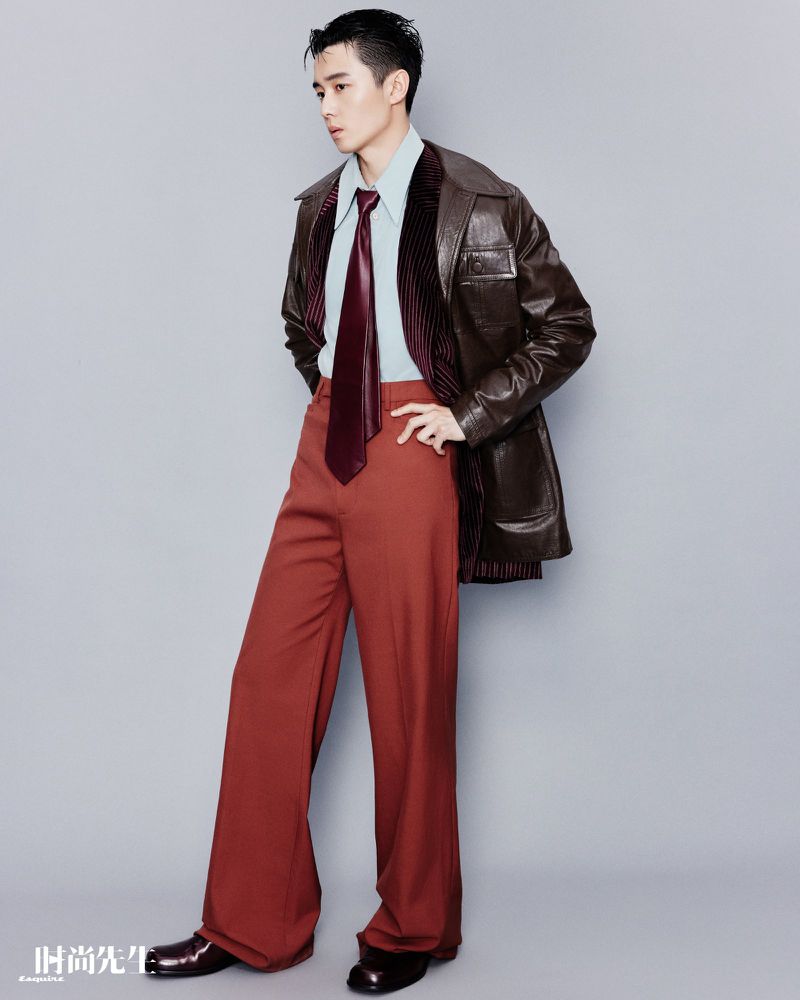 Full-length studio shot of a young adult male fashion model in brown leather jacket, teal shirt, wide tie, and flared slacks striking a pose.