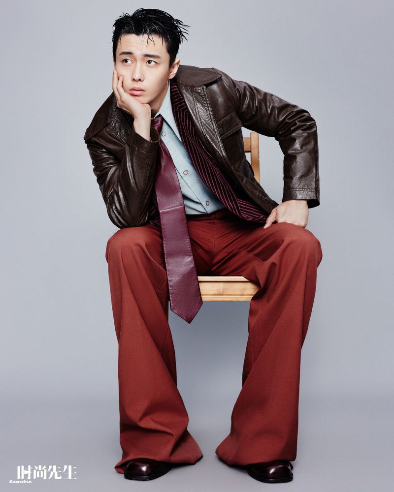 Man in brown leather jacket and red pants sits in a photo studio, looking off-camera with a serious expression, fashion editorial shot.