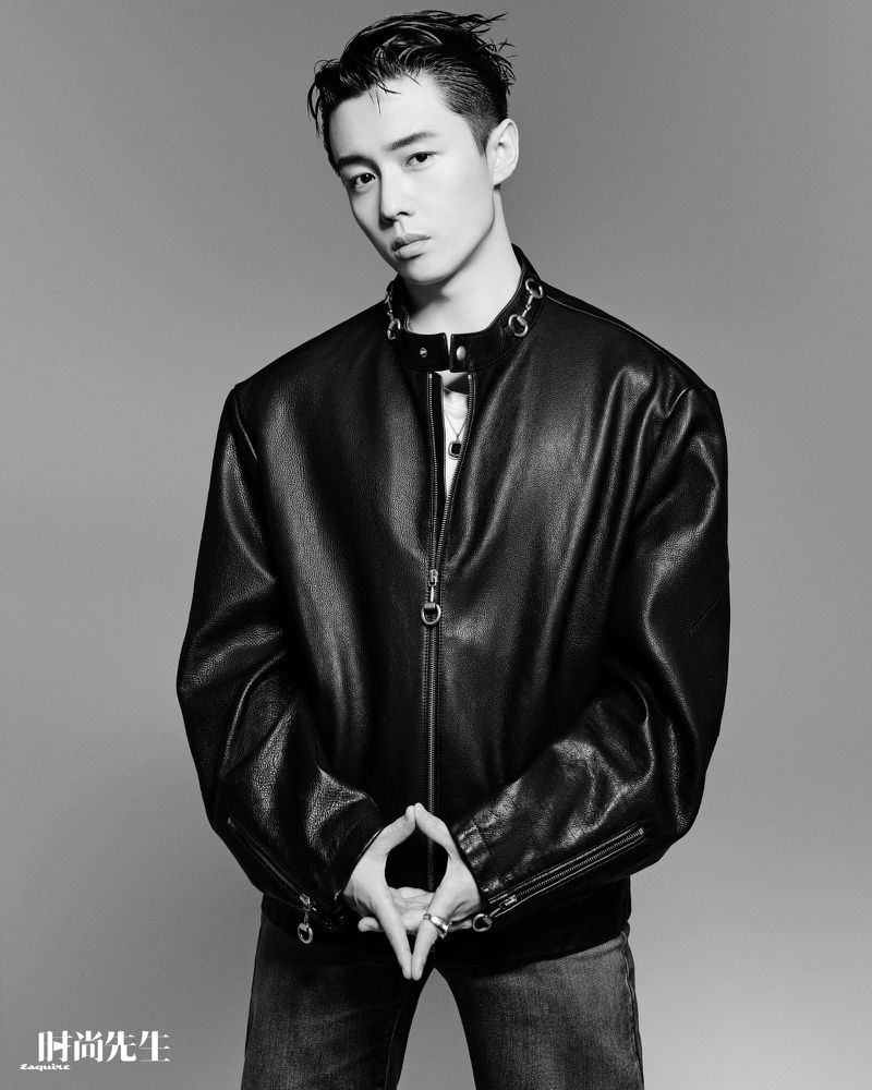 Black and white portrait of a young adult male in a leather jacket, posing with hands together and wearing denim jeans in a studio setting.