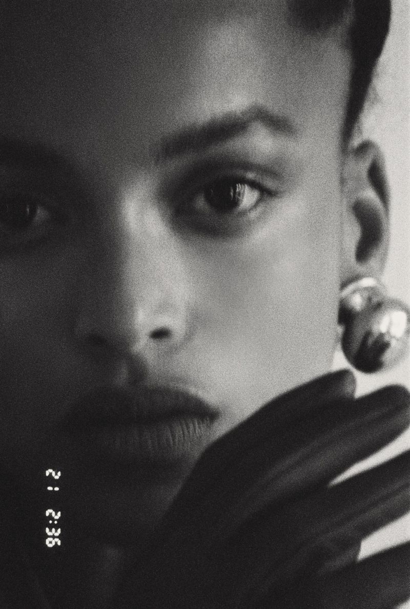 Close-up black-and-white portrait of a woman with short dark hair, wearing metallic earrings, and a dark glove covering part of her face.