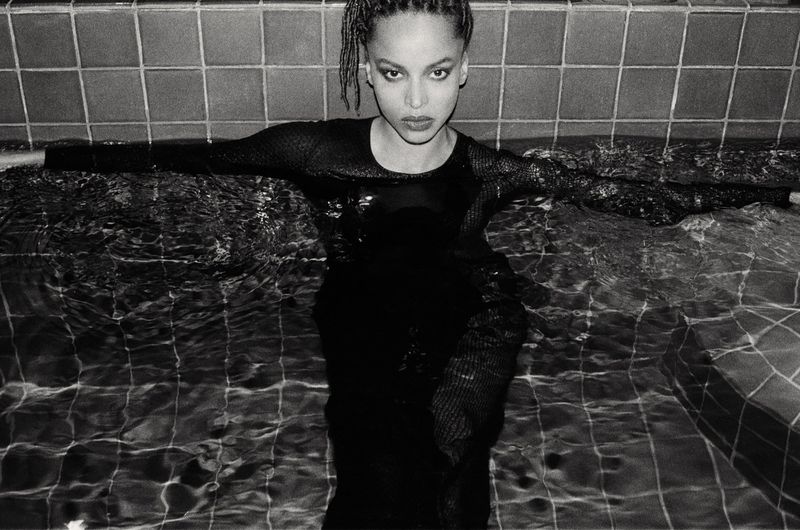 Black and white portrait of an adult woman in a dark dress standing in water with a tiled background, shot at eye-level.
