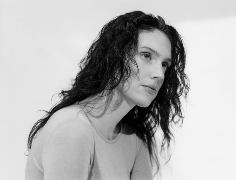 Close-up portrait of a woman with long dark wavy hair looking to the right against a plain, bright white studio background.