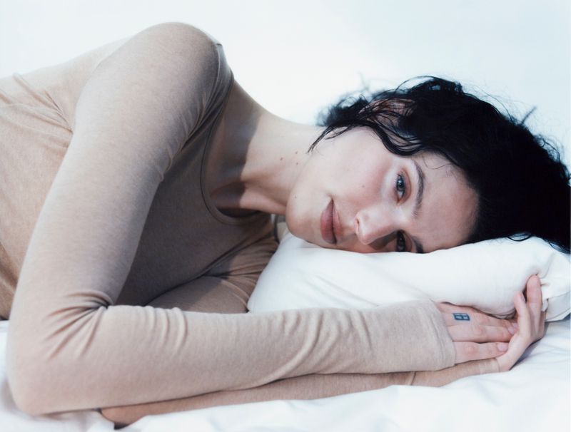 Woman with dark hair poses while lying on white bedding. She's wearing a tan long-sleeve shirt. Indoor setting, soft lighting.