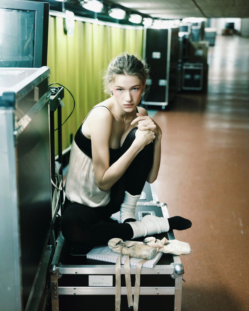 Young woman in ballet attire rests on stage equipment with ballet shoes and notes, showcasing a candid moment backstage.