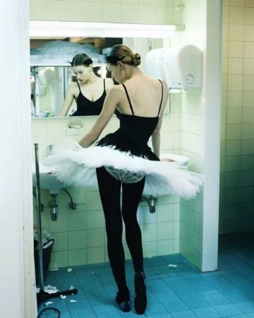 A ballerina in a black leotard and white tutu stands in a tiled bathroom, reflected in the mirror, in this indoor dance portrait.