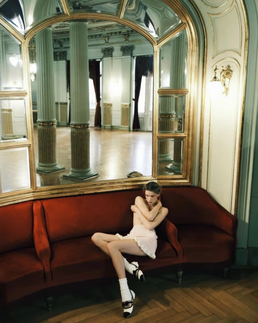 A fair-skinned young woman in a pale dress and socks sits on a red velvet couch in an elegant room reflected in a large mirror.