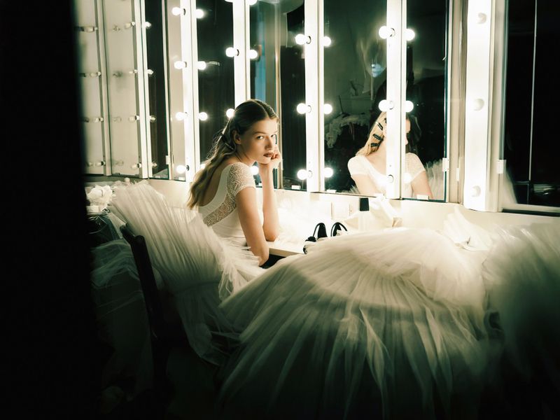 Young woman in a white dress sits in front of a lighted vanity mirror, surrounded by tulle fabric in a dressing room setting.